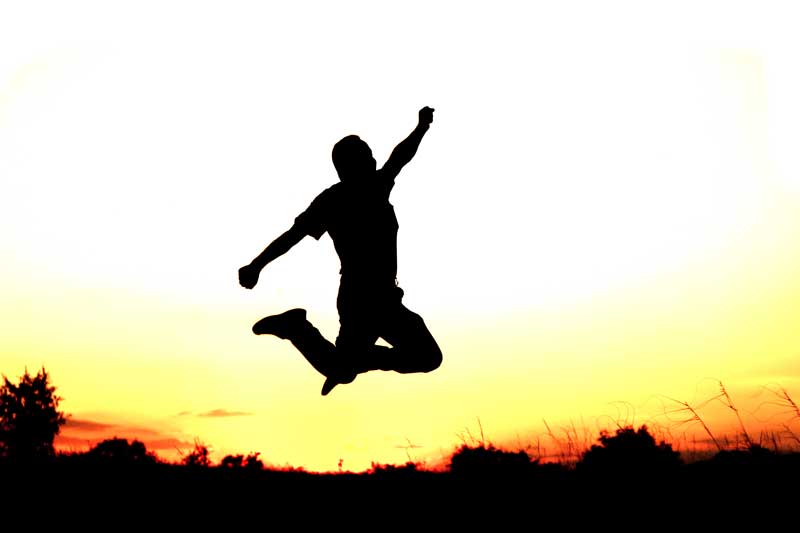 A silhouette of a man can be seen jumping triumphantly in an open field at sunset.